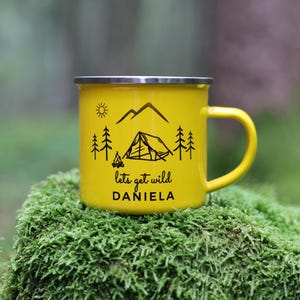 May include: Yellow enamel camping mug with a black design of mountains, trees, a tent, and a campfire. The text "lets get wild" and the name "DANIELA" are printed on the mug.