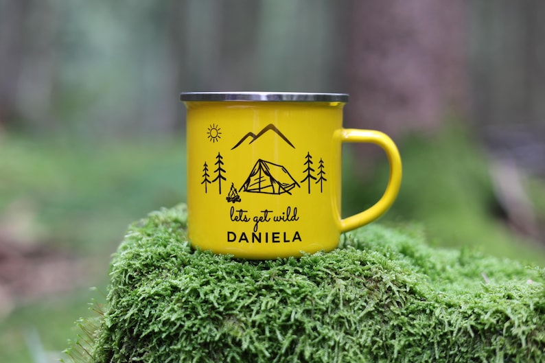 May include: Yellow enamel camping mug with a black design of mountains, trees, a tent, and a campfire. The text "lets get wild" and the name "DANIELA" are printed on the mug.