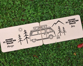 Könnte beinhalten: Zwei rechteckige Holzschilder mit Camping-Motiv. Jedes Schild zeigt eine schwarze Strichzeichnung eines Wohnmobils, Bäume, Berge, eine Sonne und ein Lagerfeuer. Der Text "DRAUBEN IST, WO WIR SEIN WOLLEN" ist auf jedem Schild zusammen mit den Namen "Monja" und "Ben" aufgedruckt.