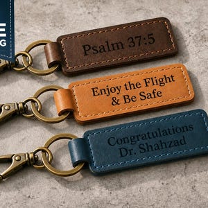 May include: Three leather keychains with metal clasps. The keychains are brown, orange, and blue. The brown keychain reads "Psalm 37:5". The orange keychain reads "Enjoy the Flight & Be Safe". The blue keychain reads "Congratulations Dr. Shahzad".