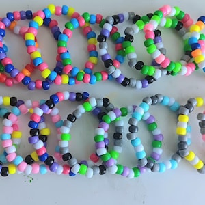 May include: A collection of colorful beaded bracelets in various color combinations. The bracelets are made of small, round beads in shades of blue, pink, yellow, green, orange, and black. The bracelets are arranged in two rows on a white surface.