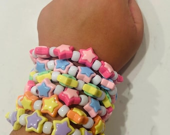 Puede incluir: Una pila de pulseras de cuentas de colores. Cada pulsera presenta cuentas en forma de estrella en tonos de rosa, amarillo, naranja, azul y morado, intercaladas con cuentas blancas. Las pulseras están apiladas en una muñeca.