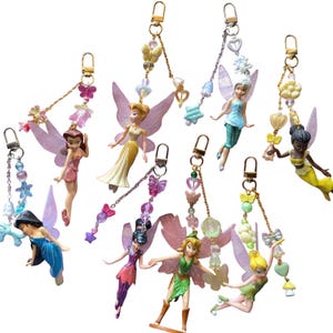 May include: Collection of fairy keychains featuring various characters with colorful wings and outfits. Each keychain has a gold-colored clasp and decorative beaded accents with butterfly and flower charms.