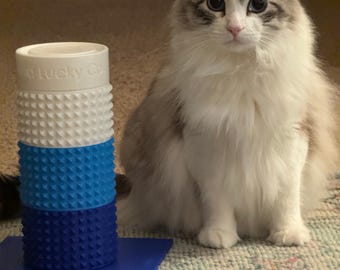 May include: A fluffy, long-haired cat with blue eyes sits next to a multi-colored cat toy. The toy is cylindrical and stacked with white, light blue, and dark blue sections. The cat is mostly white with gray markings.