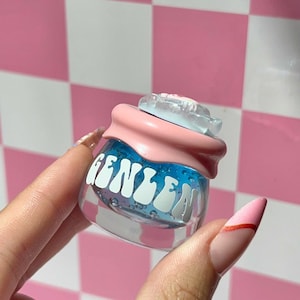 Könnte beinhalten: Ein kleines, klares Glas mit dem Wort "GENIEN" in Weiß, gefüllt mit blauer Flüssigkeit und Blasen. Das Glas hat einen rosa Deckel und eine transparente, blütenförmige Oberseite. Der Hintergrund ist ein rosa-weißes Schachbrettmuster.