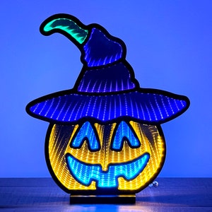 May include: A Halloween-themed LED light featuring a smiling jack-o'-lantern wearing a witch's hat. The pumpkin has a yellow and blue color scheme with a black outline. The hat is blue with a green tip. The light sits on a black base.