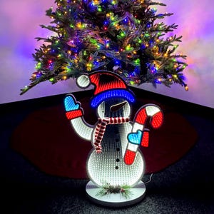 May include: A white, light-up snowman decoration with red, white, and blue accents. The snowman is holding a candy cane and has a red and white scarf. The snowman is standing on a white base with green pine branches.