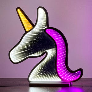 May include: A decorative unicorn-shaped LED lamp with a black outline. The unicorn has a yellow horn, a pink mane, and a white, dotted interior that creates an infinity mirror effect. The lamp sits on a wooden base and emits a soft glow.