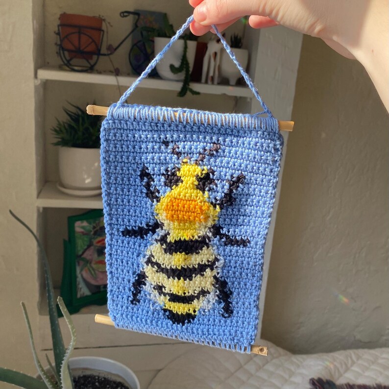 May include: A crocheted wall hanging featuring a cartoon bee with black and yellow stripes, orange and black eyes, and a light blue background.