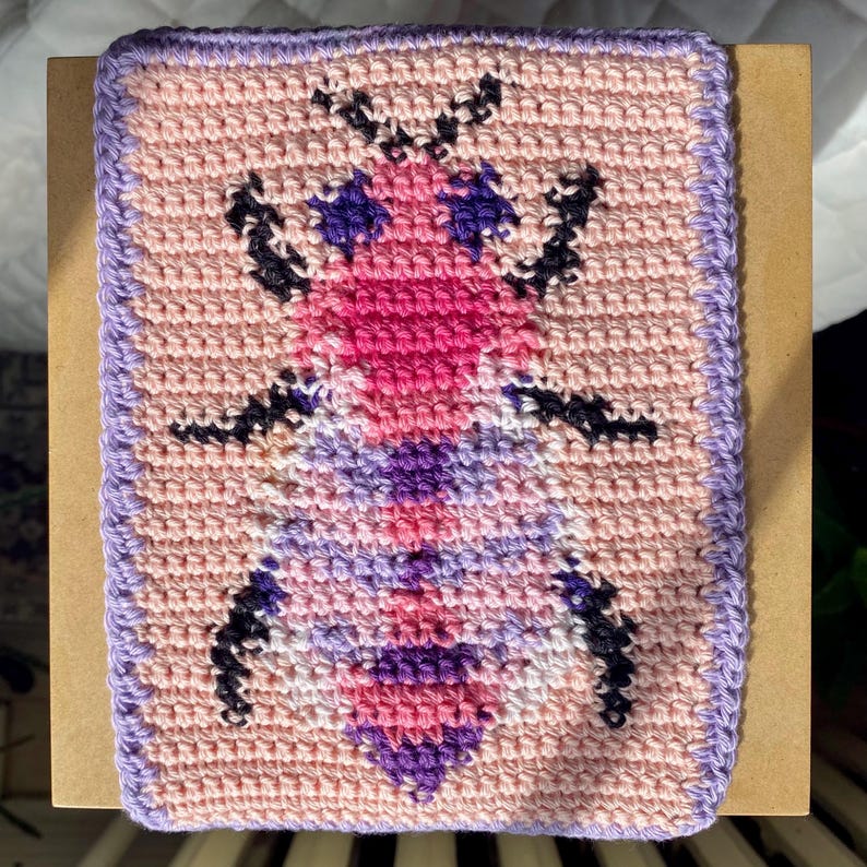 May include: A crocheted square with a pink, purple, and white bee design. The bee is outlined in black and has a pink body with purple wings.