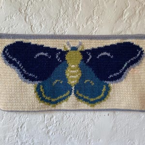 May include: A crocheted wall hanging featuring a blue and green butterfly with yellow accents. The butterfly is centered on a cream-colored background with a light blue border.