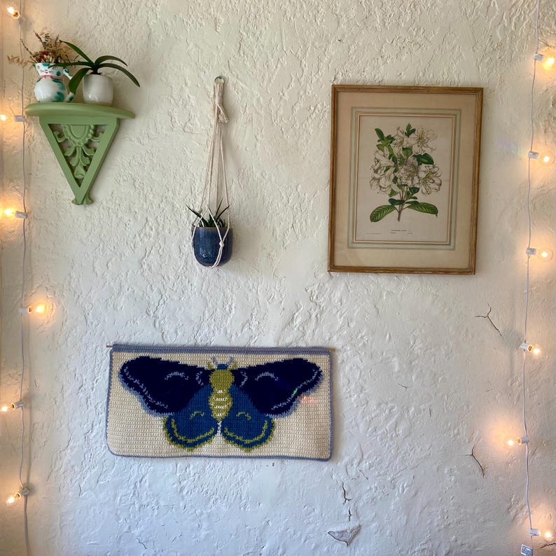 May include: A white wall with a green shelf, a framed botanical print, a hanging plant, and a blue and green butterfly needlepoint wall hanging.
