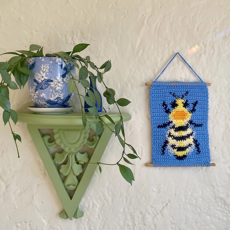 May include: A crocheted wall hanging featuring a bee with yellow and black stripes on a blue background. The hanging is attached to a wooden dowel rod.