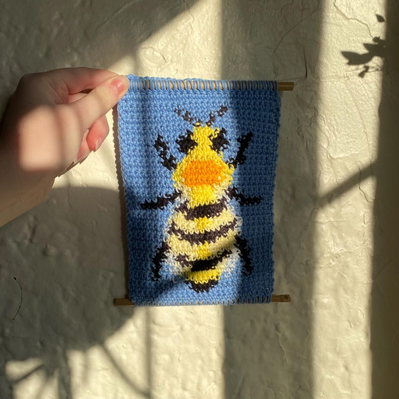 May include: A blue knitted wall hanging with a yellow and black bee design. The bee has a yellow and orange face.
