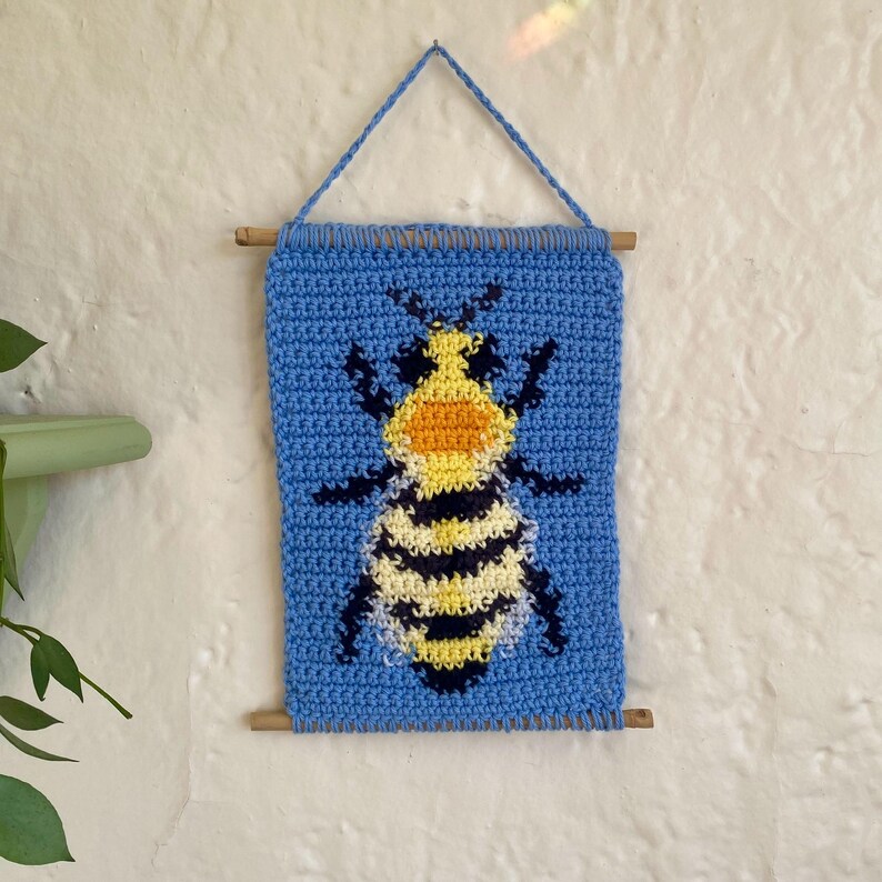 May include: A crocheted wall hanging featuring a bee with yellow and black stripes on a light blue background. The bee has a yellow and orange head and is surrounded by a blue crocheted border. The hanging is attached to a wooden dowel.