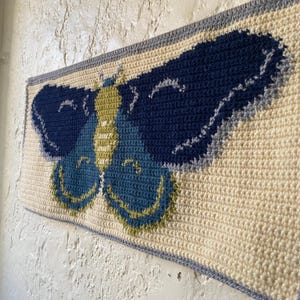 May include: A crocheted wall hanging featuring a blue and green butterfly with yellow accents. The butterfly is centered on a cream-colored background with a gray border.