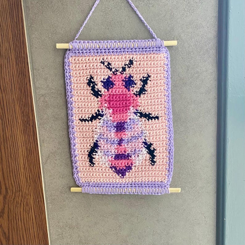 May include: A crocheted wall hanging featuring a pink and purple bee design. The bee is surrounded by a light pink background and a purple border. The hanging is attached to a wooden dowel.