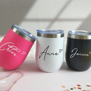 May include: Three stainless steel tumblers with clear plastic lids, each featuring a name in cursive script and a small heart icon. The tumblers are colored hot pink, white, and matte black, with the names Chloe, Anne, and Janice printed on them.