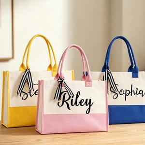 K&ouml;nnte beinhalten: Drei Canvas-Tragetaschen in Gelb, Rosa und Blau. Jede Tasche hat eine kontrastierende Bodenplatte und passende Griffe. Die Taschen sind mit Namen in schwarzer Schrift personalisiert und mit gestreiften B&auml;ndern verziert.