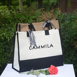 K&ouml;nnte beinhalten: Zwei Tragetaschen, eine aus nat&uuml;rlichem Jute und die andere aus wei&szlig;em Canvas mit schwarzem Boden. Auf der wei&szlig;en Tasche steht das Wort "CAMMILA" in Schwarz. Beide Taschen haben Griffe mit schwarz-wei&szlig; gestreiften B&auml;ndern. Eine rote Rose liegt davor.