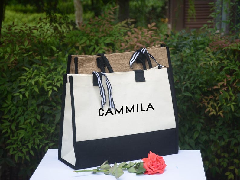 K&ouml;nnte beinhalten: Zwei Tragetaschen, eine aus nat&uuml;rlichem Jute und die andere aus wei&szlig;em Canvas mit schwarzem Boden. Auf der wei&szlig;en Tasche steht das Wort "CAMMILA" in Schwarz. Beide Taschen haben Griffe mit schwarz-wei&szlig; gestreiften B&auml;ndern. Eine rote Rose liegt davor.