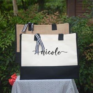 K&ouml;nnte beinhalten: Zwei Tragetaschen mit schwarzen und wei&szlig;en Akzenten. Die vordere Tasche ist wei&szlig; mit schwarzem Boden und dem Namen "Anicole" in schwarzer Schrift. Die Griffe sind schwarz, und die Tasche hat ein schwarz-wei&szlig; gestreiftes Band. Die zweite Tasche ist beige.