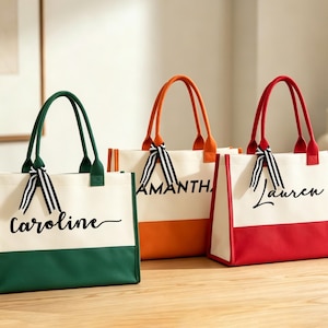 K&ouml;nnte beinhalten: Drei personalisierte Tragetaschen in Gr&uuml;n, Orange und Rot. Jede Tasche hat einen wei&szlig;en Canvas-K&ouml;rper mit farbigem Boden und passenden Griffen. Die Namen "Caroline", "Samantha" und "Lauren" sind in schwarzer Schrift auf der Vorderseite aufgedruckt, mit einem gestreiften Band an der Griff befestigt.