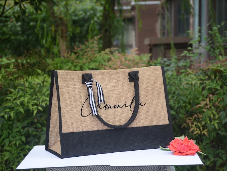 K&ouml;nnte beinhalten: Eine rechteckige Tragetasche mit nat&uuml;rlicher Jute-Textur, schwarzen Akzenten und einem schwarzen Griff. Die Tasche tr&auml;gt den Namen "Cammila" in Schreibschrift. Eine rote Rose liegt in der N&auml;he der Tasche.