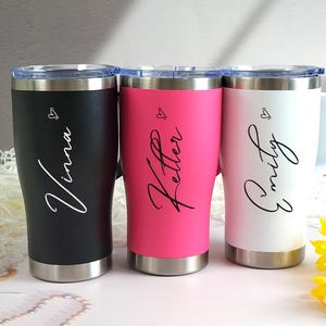 Könnte beinhalten: Drei personalisierte Becher in Schwarz, Pink und Weiß, jeweils mit silberfarbenem Boden und transparentem Deckel. Die Becher zeigen Namen in eleganter Schrift: Vinna, Kettler und Emily, zusammen mit einem kleinen Herzdesign. Die Becher haben Griffe.