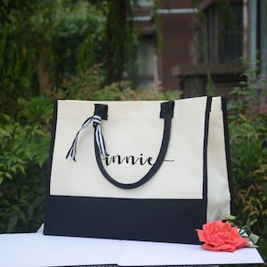 K&ouml;nnte beinhalten: Eine cremefarbene und schwarze Canvas-Tragetasche mit schwarzen Griffen und Verzierungen. Der Name "innie" ist in Schreibschrift auf die Tasche geschrieben. Ein gestreiftes Band ist an einem Griff befestigt. Eine Korallenrose liegt vor der Tasche.