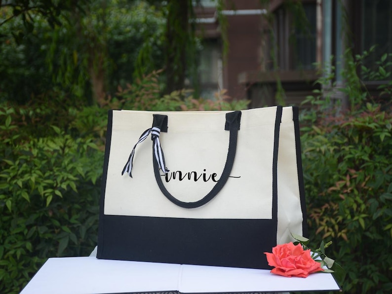K&ouml;nnte beinhalten: Eine cremefarbene und schwarze Canvas-Tragetasche mit schwarzen Griffen und Verzierungen. Der Name "innie" ist in Schreibschrift auf die Tasche geschrieben. Ein gestreiftes Band ist an einem Griff befestigt. Eine Korallenrose liegt vor der Tasche.