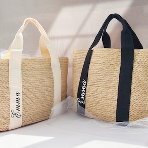 May include: Two straw tote bags with black and white straps. The bags are personalised with the name "Emma" in white lettering.