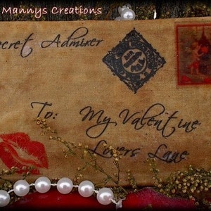 Op de afbeelding: Een verouderde envelop met de woorden "Secret Admirer" en "To: My Valentine Lovers Lane" in elegant schrift. Voorzien van een rode kusafdruk, een decoratieve postzegel en een zwart ruitvormig ontwerp. Versierd met parels en gedroogde bloemen.