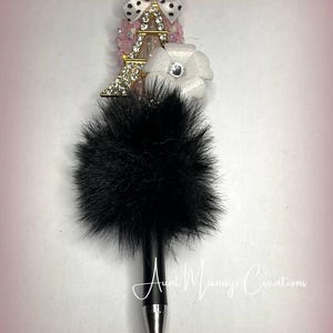 May include: Black and white pom pom pen with a gold Eiffel Tower charm, a white flower charm with a rhinestone center, and a black and white polka dot bow.