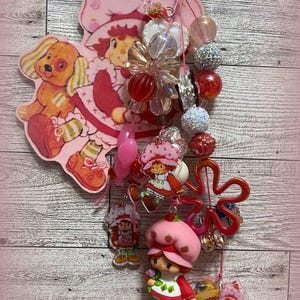 Może przedstawiać: Kolekcja zawieszek i koralików z motywem Strawberry Shortcake. Zawieszki zawierają wizerunki Strawberry Shortcake i jej przyjaciół, a także psa. Koraliki są w różnych kolorach i kształtach, w tym przezroczyste, czerwone i opalizujące. Widoczny jest tekst "Aunt Manny's Creations".