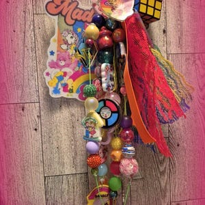 May include: A decorative keychain featuring a large acrylic charm with cartoon characters and the word Mad, a miniature Rubik's cube, various colorful plastic beads, a circular game-themed charm, and layered ribbons in bright orange, pink, and yellow.