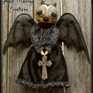Pode incluir: Uma boneca de morcego preta com grandes olhos verdes e um vestido preto segurando uma cruz cinza. A boneca está em pé contra um fundo de madeira. O texto "Aunt Mannys Creations" é visível no canto superior esquerdo da imagem.