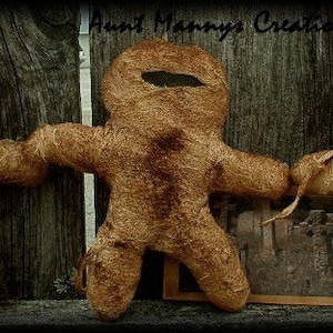May include: Three brown burlap dolls with black eyes and mouths, wearing burlap hoods. The dolls are arranged in a row on a wooden surface. The text "Aunt Mannys Creations" is visible in the background.