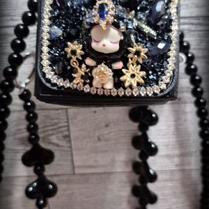 May include: A black purse with a small, white doll with black hair and a blue crown. The purse is decorated with rhinestones, gold stars, and black beads. The purse is attached to a black beaded chain.