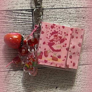 May include: A keychain featuring a red strawberry charm, a clear acrylic charm with a Strawberry Shortcake character, and a small pink notepad with a Strawberry Shortcake design. The keychain is attached to a silver clasp.