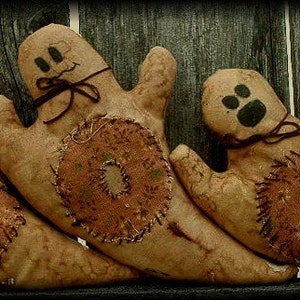 May include: Three handmade ghost decorations with stitched details and smiling faces. Each ghost has a unique patch on its body, and they are tied with a dark brown ribbon. The ghosts are made of tan fabric and are arranged against a dark wooden background.