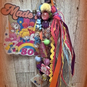 Op de afbeelding: Een decoratieve hangende bedel met een groot uitgesneden bord met de tekst Made in the 80s, kleurrijke stripfiguren en een regenboog. Het bevat een cluster van veelkleurige kralen, kleine bedels en een bundel van diverse stoffen linten.