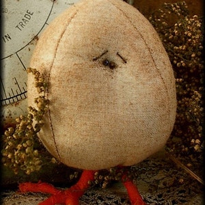 May include: A handmade fabric chick with a beige body, red legs, and a button eye. The chick is sitting on a lace doily and surrounded by dried flowers.