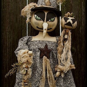 Op de afbeelding: Een handgemaakte heksenpop met een zwart-wit gebloemde jurk, een punthoed en een pompoen in haar hand. De pop is gemaakt van stof en heeft een rustieke, vintage uitstraling. De pop staat op een houten oppervlak. "Aunt Mammys Creations" staat op de achtergrond.