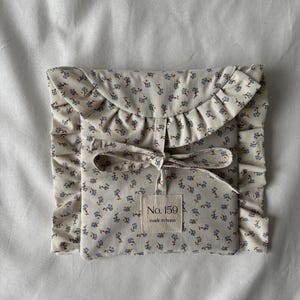 Könnte beinhalten: Eine cremefarbene Tasche mit floralem Muster, Rüschenkante und Schleifenverschluss. Die Tasche hat kleine blaue Blumen und ein Etikett mit der Aufschrift "No. 159 made in bonn."