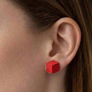 Könnte beinhalten: Nahaufnahme eines Ohrs mit einem leuchtend roten, würfelförmigen Ohrstecker. Der Ohrring hat eine einfache geometrische Form. Die Haut um das Ohr ist hell, und braunes Haar ist sichtbar.