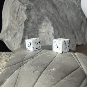 Op de afbeelding: Twee kleine, vierkante, witte dobbelstenen met zwarte marmering liggen op een grijs oppervlak. De dobbelstenen zijn scherp gefocust, met een grote, grijze rotsformatie op de achtergrond. De esthetiek is minimalistisch en modern.