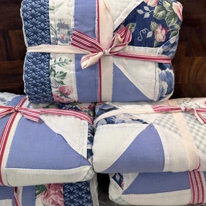 May include: Stacked, folded quilts tied with pink and red striped ribbon. The quilts feature a patchwork design with floral, blue patterned, and solid light blue fabrics. The edges are white.