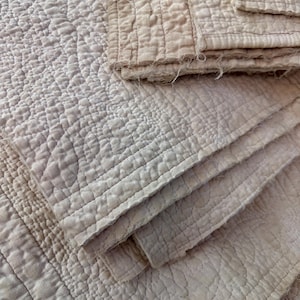 May include: Close-up of a light beige quilted fabric. The fabric has a textured, raised pattern with stitched lines and a folded edge. The overall appearance is soft and vintage.