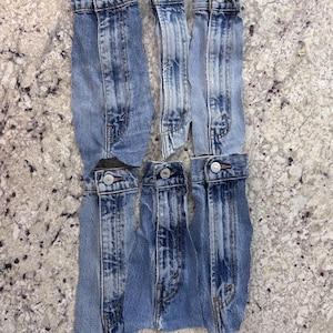 May include: Five detached blue denim jean waistbands arranged in two rows. Each waistband features a silver-toned metal button closure, visible belt loops, and frayed edges where the fabric has been cut from the original garment.